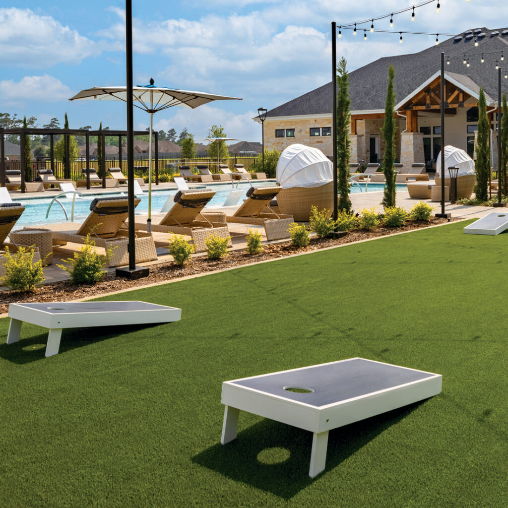 Outdoor photo of greenery,turf, cornhole, cabanas, string lights, lounge chairs and pool with tanning ledges.