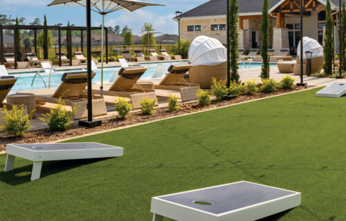 Outdoor photo of greenery,turf, cornhole, cabanas, string lights, lounge chairs and pool with tanning ledges.